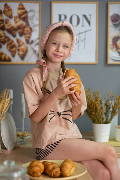 a girl in beige pajamas and a pink headband and with pigtails sits on the kitchen table and eats a croissant. children's nutrition. upbringing. the child is getting ready for bed. child protection. children's Day. child psychology.