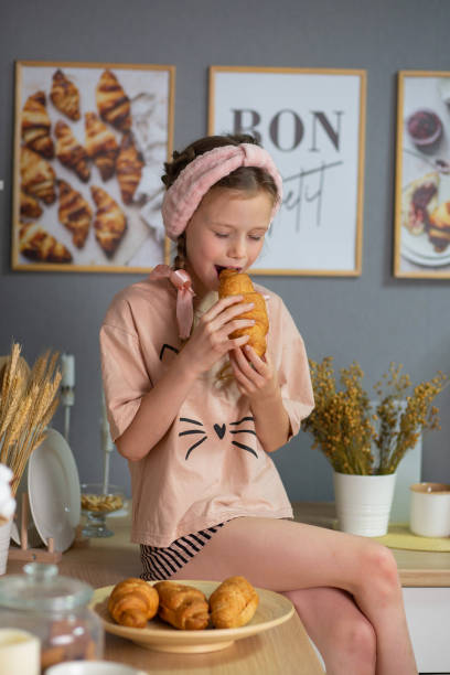 a girl in beige pajamas and a pink headband and with pigtails sits on the kitchen table and eats a croissant. children's nutrition. upbringing. the child is getting ready for bed. child protection. children's Day. child psychology.