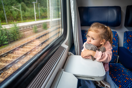 Bored little girl with toy looking out train window outside, while it moving. Traveling by railway, Europe. High quality photo