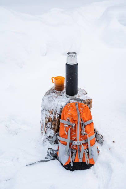 istock Black matte thermos, orange backpack for hiking and a mug with tea on a snow-covered stump. Drink warm tea on a hike in winter forest. Minimalistic subject shot 1759257516