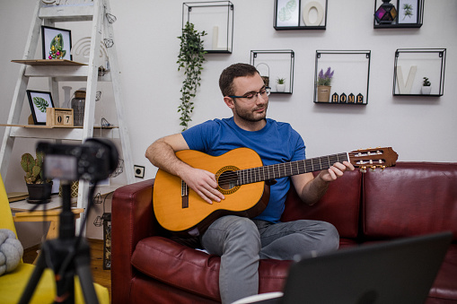A Caucasian adult male learning to play the guitar through online classes on his laptop in his apartment