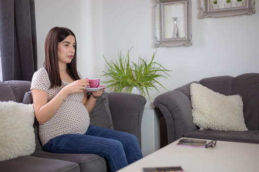 The pregnant woman is sitting on the sofa and holding a small , pink cup of tea. The woman is relaxing at home.