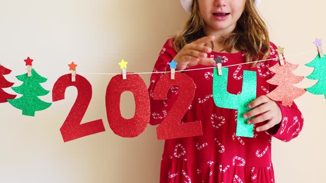 Child girl in red Christmas dress decorates home for holidays New Year party of sparkling handmade garland, kid hanging change glitter numbers 2023 to 2024. crafts for kids DIY. decoration concept