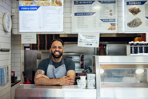 A medium shot of a mid-adult Indian man wearing casual clothing and an apron. He is standing behind the counter of his family's fish and chips shop. He looks and smiles at the camera.