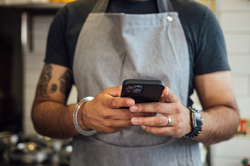 A close-up shot of an unrecognisable mid-adult Indian man wearing casual clothing and an apron. He is standing behind the counter of his family's fish and chips shop. He holds his smartphone in both hands.