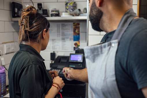 A medium, over-the-shoulder shot of a mid-adult Indian couple wearing casual clothing and aprons. They work together in the back of their family-run fish and chip shop.