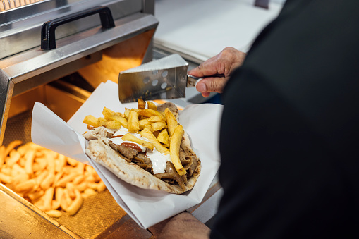 An over-the-shoulder, medium shot of an unrecognisable senior Indian man wearing casual clothing. He is working in his family-run fish and chip shop. He makes up a wrap filled with kebab meat, chips, garlic and chilli sauce.