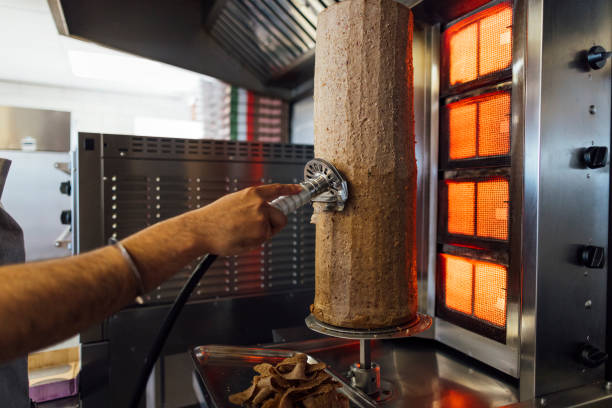 A medium shot of an unrecognisable mid-adult Indian man wearing casual clothing and an apron. He is working in his family-run fish and chip shop. He uses a kebab shaver to cut the doner kebab.