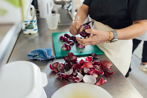 A medium shot of an unrecognisable senior Indian woman wearing casual clothing and an apron. She is working in her family-run fish and chip shop. She cuts a red onion as she prepares food for the day ahead.