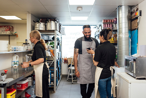 A medium shot of a mid-adult Indian couple and their mother wearing casual clothing and aprons. They work together in the back of their family-run fish and chip shop.