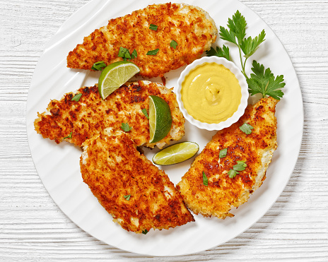 fried chicken cutlets under breadcrumbs and parmesan cheese crust on white plate with mustard and lime on white wooden table, horizontal view from above, flat lay, close-up
