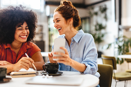 Two female colleagues enjoying a coffee break in their coworking space, reading a client’s text message on a smartphone. Diverse business women smiling as they work together.