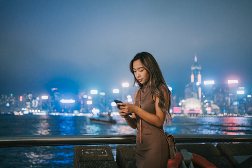 Young Asian woman using smartphone by the promenade Star Avenue at night, with illuminated and multi-coloured urban city skyline in background. Lifestyle and technology