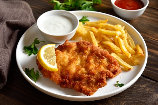 Breaded wiener schnitzel with potato fries and sauce on wooden table. Close up vuew