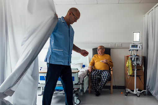 A senior male patient with cancer staying in a hospital in Newcastle upon Tyne, North East England. A male staff nurse is pulling a curtain around the area the patient is in to create some privacy so he can perform medical tests. The patient is sitting on a chair next to his hospital bed ready to receive the tests.