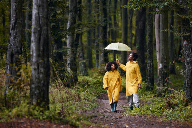 coppia nera felice in impermeabili che cammina sulla pioggia attraverso i boschi. - candid women african descent umbrella foto e immagini stock