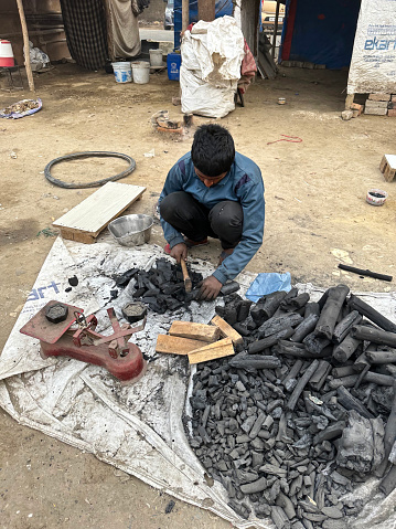 Noida, Uttar Pradesh, India - January 09, 2023: Stock photo showing close-up, elevated view of a heap of natural wood charcoal being chopped up with an axe by a vendor with an axe in order to weigh and sell to customers.