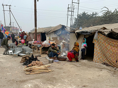Noida, Uttar Pradesh, India - January 09, 2023: Stock photo showing people sitting before makeshift shelters with tree branches and tarpaulin covers and sheds, slum set up on waste ground.