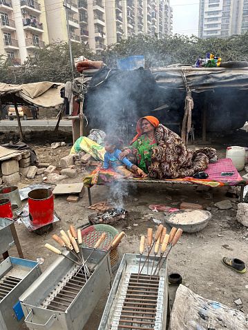 Noida, Uttar Pradesh, India - January 09, 2023: Stock photo showing family sitting on cot bed before makeshift shelters with tree branches and tarpaulin covers, slum set up on waste ground with background of residential tower blocks.
