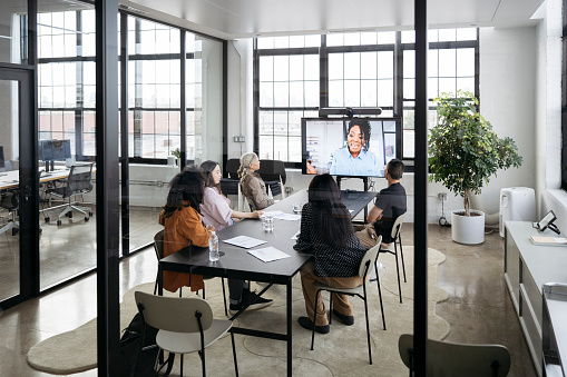 Full length view of professionals sitting at conference table in modern see-through meeting room and listening to progress report.
