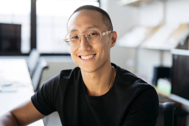 Desk portrait of confident young Taiwanese businessman Head and shoulders view of shorthaired professional seated at workstation in modern office, wearing black t-shirt and eyeglasses, smiling at camera. taiwan people stock pictures, royalty-free photos & images