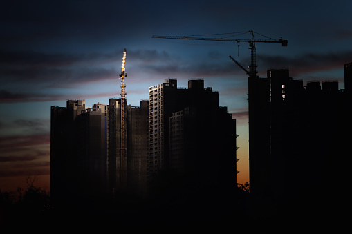 Tower cranes at new city building and apartment construction sites, red sunset sky and clouds, dark night building silhouettes, and construction industry and real estate development background
