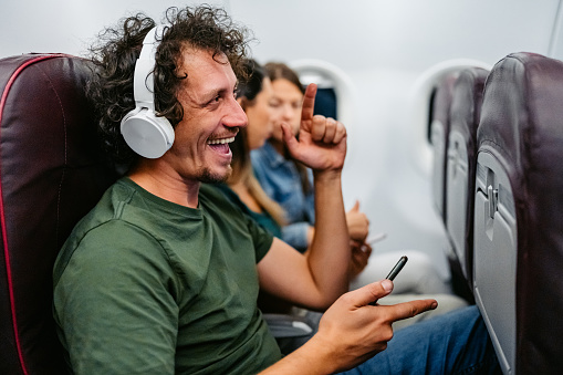 Handsome young man listening to music via headphones using his smart phone in an airplane.