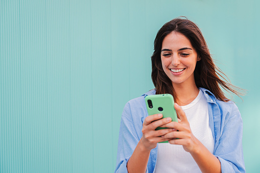 Smiling happy teenage woman using her smartphone and chatting online ,or browsing on internet at blue teal wall background. Young caucasian college student lady watching funny videos with mobile phone. High quality photo