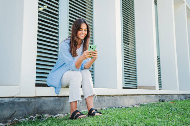 istock joyful caucasian woman smiling and browsing on internet with a mobile phone, texting emails, connecting people sitting outside. Young teenage female student using a cellphone app at university campus 1759457931