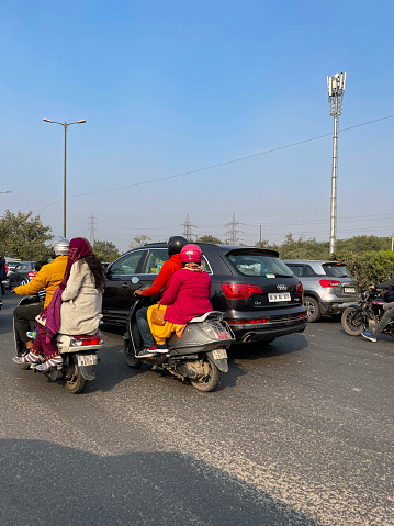 Greater Noida near New Delhi, India - January 1, 2023: Stock photo showing close-up view of busy road in Greater Noida near New Delhi congested with traffic including cars, motorbikes and mopeds carrying pillion passengers with no safety helmets behind motorcyclist with crash helmets.