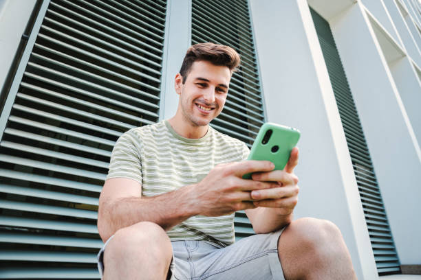 istock Young guy with friendly expression smiling and having fun playing a video game with his cellphone sitting outdoors. Caucasian teenage stundent enjoying using a mobile phone for watch the social media 1759469504