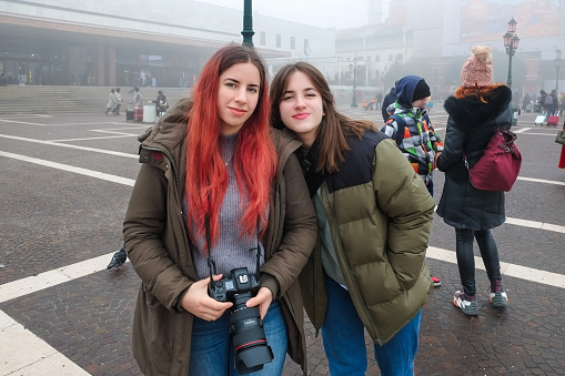 Beautiful young woman and her teenage sister standing in town square on a foggy day and looking at camera, visiting Italy together at wintertime