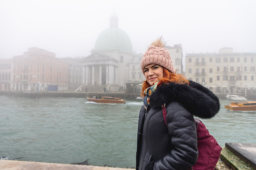 Beautiful woman in warm clothing standing by Grand Canal, looking at camera with a smile, foggy day in winter, visiting Venice in Italy