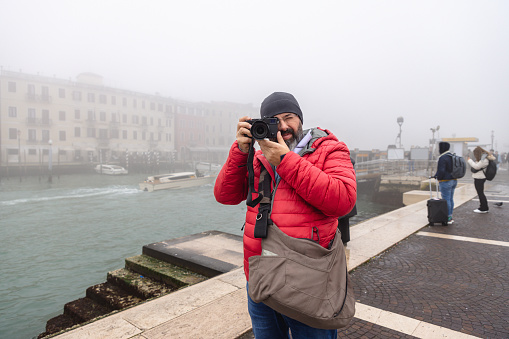 Portrait of middle aged man in warm clothing standing by Grand Canal and taking a photo towards camera, foggy day in winter, visiting Venice in Italy