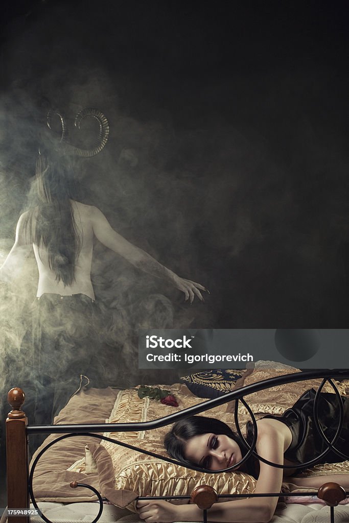 Devil from dreams Innocent gothic girl sleeps in bed. Horned horrible devil stands behind the bed in smoke. Halloween Stock Photo