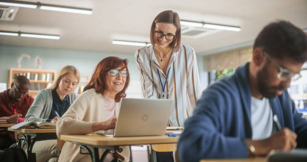 istock Helpful Young Teacher Talking with a Senior Woman, Helping to Find a Solution for a Test Course Assignment. Multiethnic Women and Men of Different Age Acquiring New Academic Skills in Classroom 1759516467