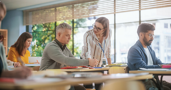 Young Female Teacher Giving a Lecture During an Adult Education Course in School, Having a Conversation with a Middle Aged Man. Diverse Mature Students Doing Textbook Exercises in Classroom