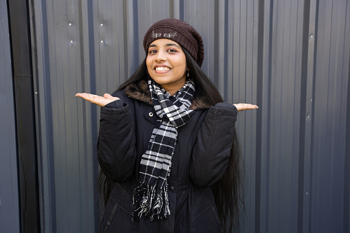Young indian happy woman wearing winter jacket standing with open hands, warm scarf and cap. isolated over grey background.