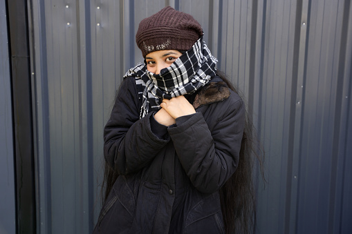 Young indian woman wearing winter jacket with warm scarf on face and cap standing isolated over grey background.