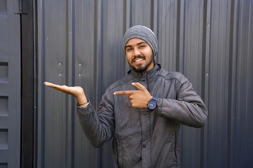young indian men wearing warm winter clothes pointing to the side to present product. Standing isolated over black background