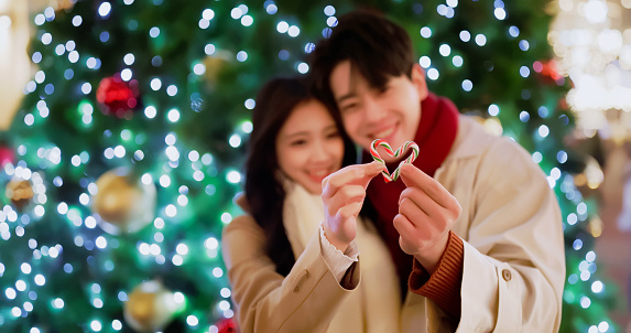 asian dating couple wearing scarves are standing in front of xmas tree and using candy cane to shaped a heart sign