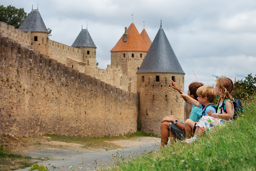 Three kids on vacations pointing at towers, having rest near walls of famous Carcassonne French fortified city in Occitania, France