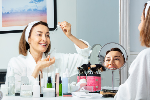 Beautiful middle aged woman putting facial serum with eyedropper on the face, wide smiling before make in mirror