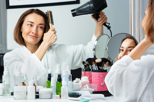 Beautiful middle aged woman in housecoat holding hairdryer dry hair before makeup mirror