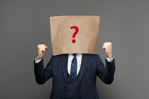 Corporate businessman hiding under paper bag with question mark on grey background