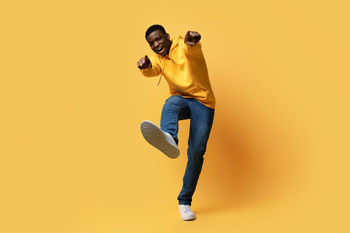 Happy handsome curly young black man in stylish casual outfit showing thumb ups and shoe sneaker sole on yellow studio background, like something or someone, copy space. Human gestures concept