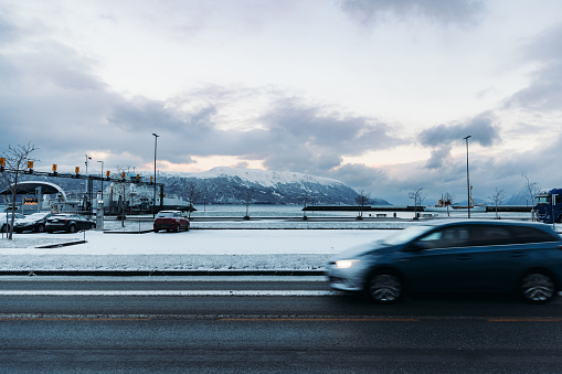 Scenic view of the cars driving the frozen road near the harbour with a ferry boat and the view of snowcapped mountains during sunset in More og Romsdal, Scandinavia