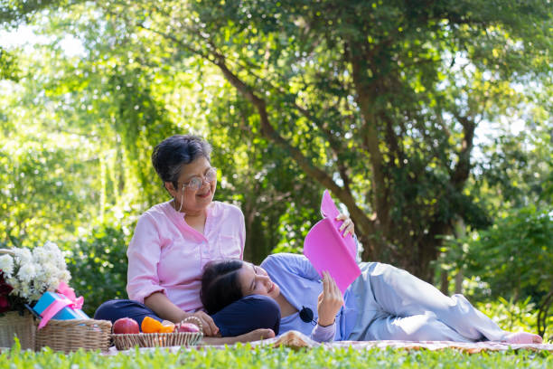 アジアの娘 母親の膝の上で眠り、公園のピクニックマットの上で本を読んでいます。幸せそうな先輩女性が娘と話す。医療・介護支援サービスのコンセプト - grandparent senior adult child reading ストックフォトと画像
