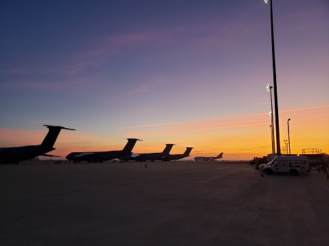 A plane is seen parked on the tarmac at sunset, with two large buildings in the background
