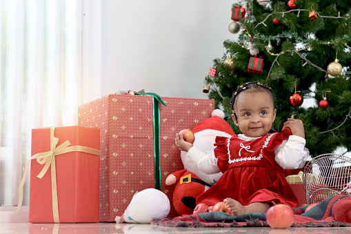 Portrait of African little girl child sitting on floor at living room with many present gift boxes under beautiful decorative Christmas tree. Cute kid celebrating happy Christmas winter holiday.